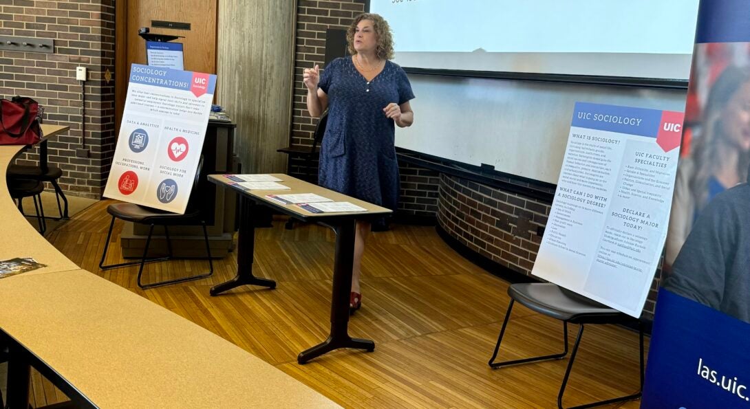 Dr. Pamela Popielarz, an adult woman with shoulder length curly hair and wearing a blue dress, is shown giving a lecture to a large lecture hall.